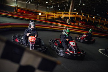 interracial men driving go kart near checkered black and white racing flag on blurred foreground clipart