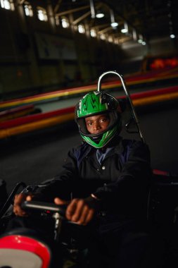 african american go kart driver in helmet driving on indoor circuit, speed racing track, motorsport clipart