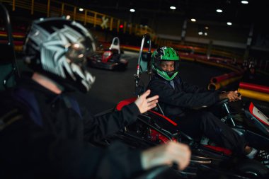 african american go kart driver in helmet looking at friend and  driving on circuit, competition clipart