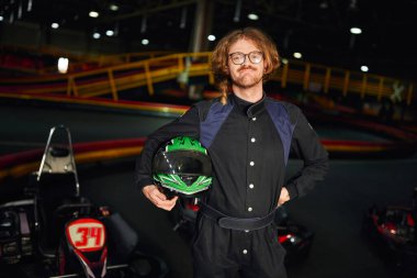 Kart sürücüsü gözlüklü ve spor kıyafetli içerde yarış pistinin kaskıyla ayakta duruyor.