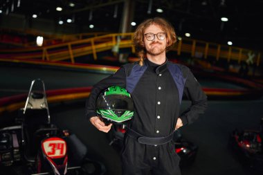 go kart driver in glasses and sportswear standing with helmet inside of indoor racing track clipart