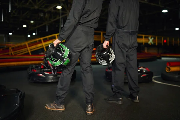 cropped view of two men standing near racing cars and holding helmets, go-kart drivers team