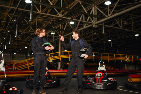kart racer pointing at friend in glasses and standing near racing cars while holding helmets