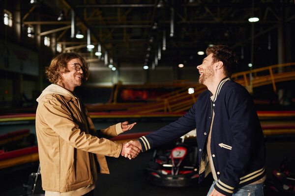 happy man in glasses shaking hands with excited friend inside of karting racing track, competition