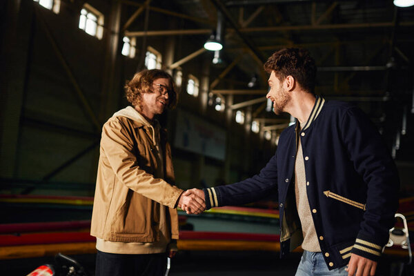 happy man in glasses shaking hands with joyful friend inside of karting racing track, competition