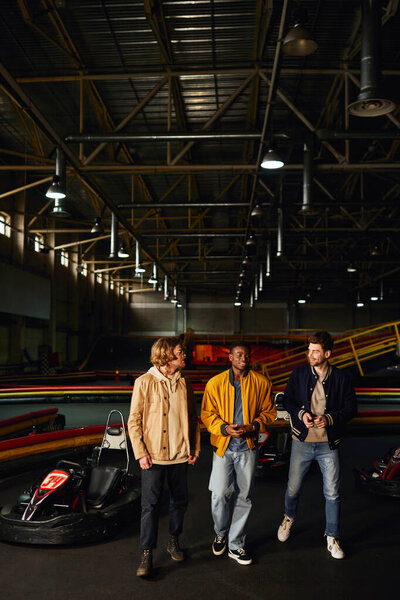 cheerful diverse friends chatting and walking near racing cars inside of karting track, motorsport