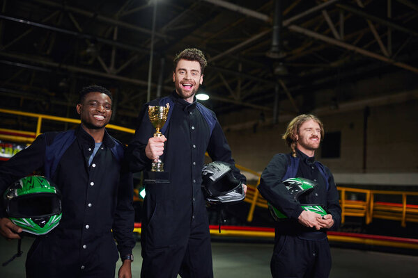 excited driver holding golden cup and standing as a winner of go-kart race near diverse competitors
