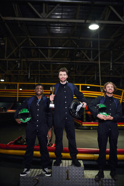 счастливый человек, держащий золотой кубок и стоящий в качестве победителя картинговой гонки вблизи межрасовых конкурентов