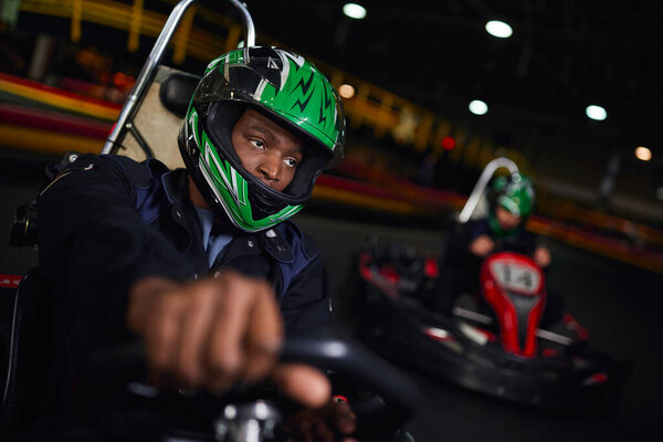 african american man in helmet driving go kart on indoor circuit near friend on blurred backdrop
