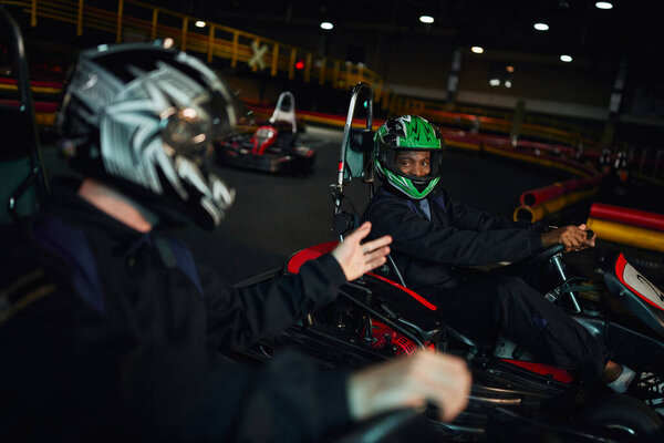 african american go kart driver in helmet looking at friend and  driving on circuit, competition