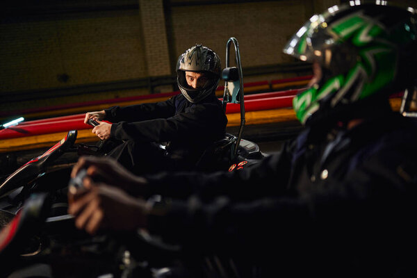 go kart driver in helmet looking at african american friend and  driving on circuit, competition