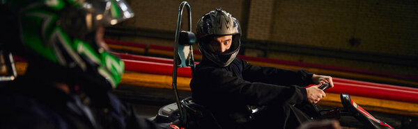 go kart driver in helmet looking at african american friend and  driving on circuit, banner