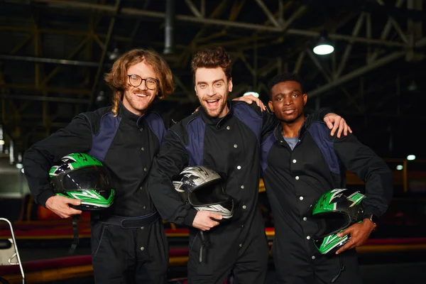 stock image multicultural and excited go kart racers in protective suits hugging and holding helmets, winners