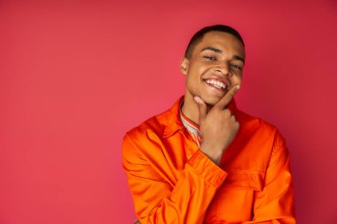 happy african american man in orange shirt touching face and looking away on red background clipart