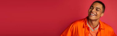 Heyecanlı Afrikalı Amerikalı adam kırmızı arka planda gülüyor, turuncu gömlek, portre, afiş, kopya alanı