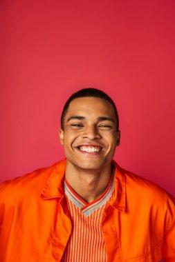 İyimser Afrikalı Amerikalı adam kırmızı arka planda kameraya gülümsüyor, turuncu gömlek, portre