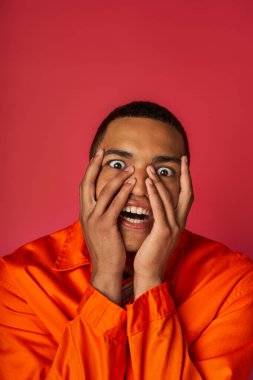 scared african american man in orange shirt obscuring face with hands and looking at camera on red clipart
