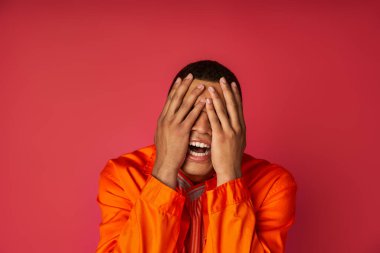 young depressed african american man in orange shirt obscuring face with hands on red background clipart