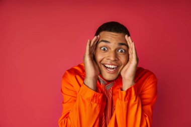 overjoyed african american man in orange shirt, with hands near face looking at camera on red clipart