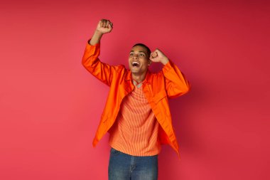 Heyecanlı turuncu tişörtlü Afro-Amerikalı adam kırmızı arka planda başarı işareti yapıyor ve bağırıyor.