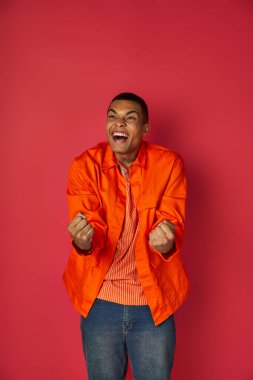 delighted, excited african american shouting and showing win gesture, orange shirt, red background clipart
