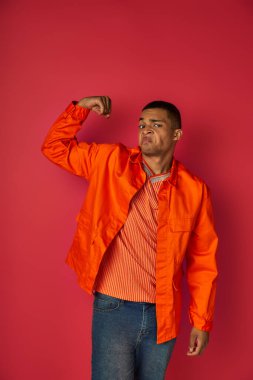 haughty african american guy demonstrating strength, looking at camera, orange shirt, red background clipart
