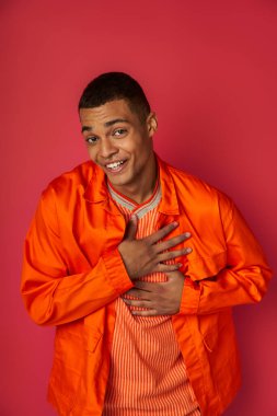 grateful and smiling african american man in orange shirt touching chest, looking at camera on red clipart