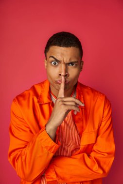 strict african american man in orange shirt looking at camera and showing hush gesture on red clipart
