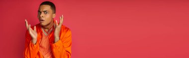 shocked african american in orange shirt gesturing and looking at camera on red, banner, copy space clipart