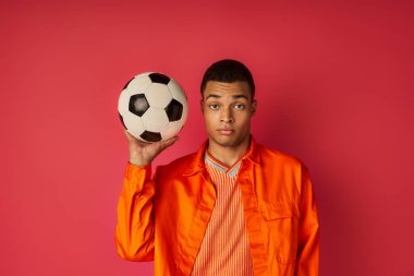Turuncu tişörtlü, futbol topu tutan ve kırmızı ışıkta kameraya bakan Afro-Amerikan genç adam.