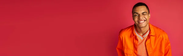 赤い背景 バナー コピースペースでカメラでウィンクするオレンジ色のシャツのアフリカ系アメリカ人の笑顔 — ストック写真