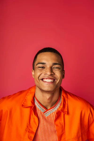 楽観的なアフリカ系アメリカ人男性 赤い背景のカメラで笑顔 オレンジ色のシャツ 肖像画 — ストック写真