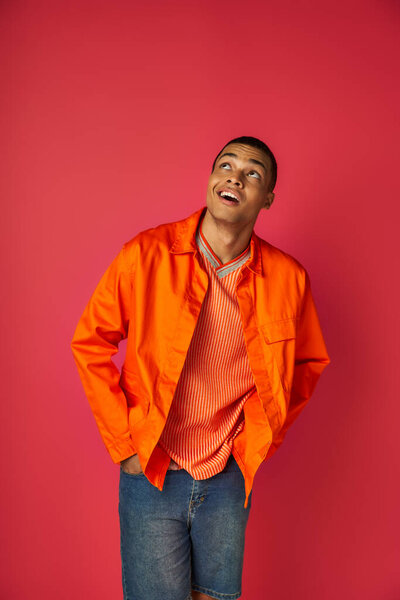 positive and curious african american in orange shirt looking up on red background, hands in pockets
