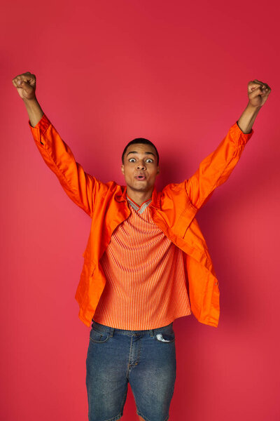 overjoyed african american man in stylish orange shirt showing win gesture on red background