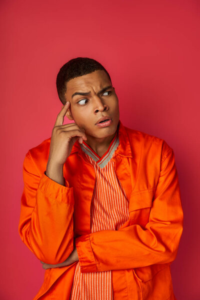 worried and tense african american man in orange shirt looking away on red background