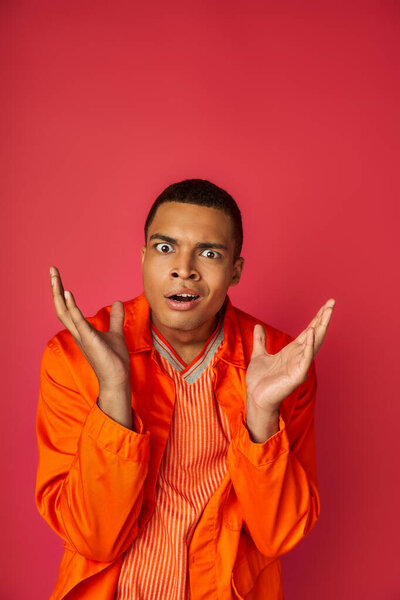 discouraged african american man in orange shirt gesturing and looking at camera on red