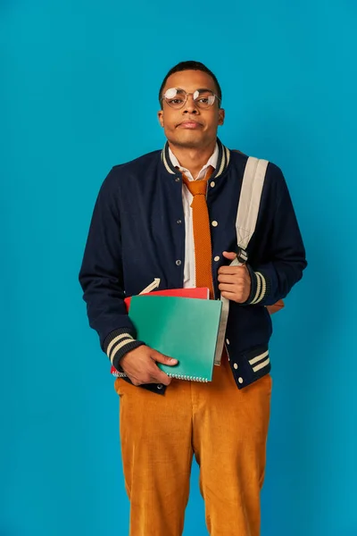 tired african american student with notebooks and backpack puffing cheeks, looking at camera on blue