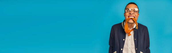 модный африканский студент в стильной одежде кусая галстук, веселясь на голубом, баннер