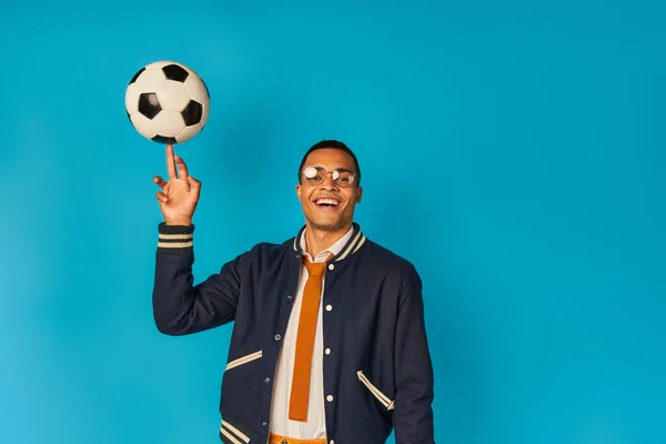 cheerful and stylish african american student playing with soccer ball and looking at camera on blue