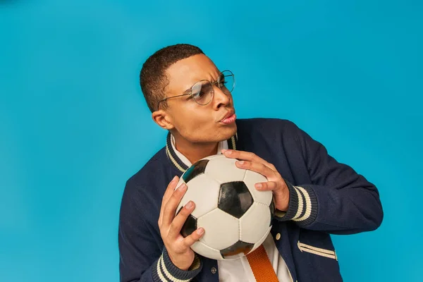 stock image serious african american student in eyeglasses holding soccer ball and looking away on blue
