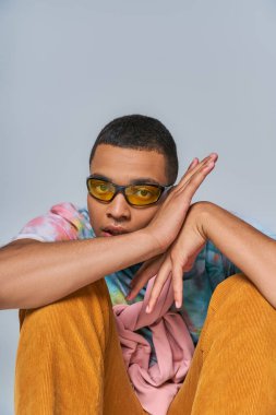 trendy african american guy in trendy sunglasses and tie-dye t-shirt looking at camera on grey clipart