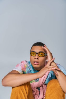 Moda gözlüklü, kravat boyalı tişörtlü, gri renkli kameraya bakan Afro-Amerikalı genç bir adam.