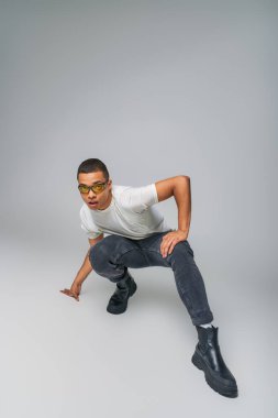 Modern tarz Afro-Amerikalı tişörtlü, kot pantolonlu ve güneş gözlüklü adam gri kameraya bakıyor.