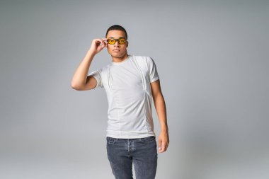 Moda trendi, Afro-Amerikalı tişörtlü, kot pantolonlu ve güneş gözlüklü adam gri kameraya bakıyor.