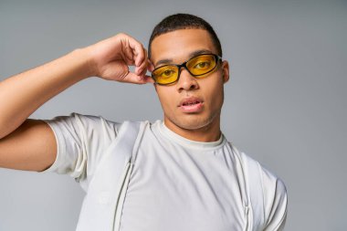Genç, şık Afro-Amerikan tişörtlü adam güneş gözlüğünü ayarlıyor, gri kameraya bakıyor.