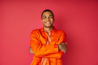 happy african american man in orange shirt, with folded hands, looking at camera on red clipart