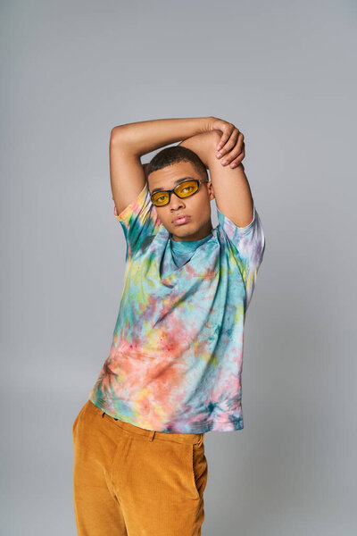 stylish african american guy in sunglasses and tie-dye t-shirt with hands above head on grey