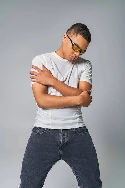 young african american man in stylish t-shirt, jeans and sunglasses with folded arms on grey