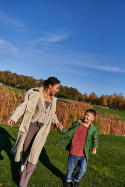 bonding, autumnal nature, happy african american mother holding hands with son, family in outerwear clipart