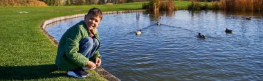 happy african american boy in outerwear sitting near pond with ducks, nature and kid, banner clipart